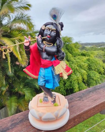 RadhaVallabha deity of Krishna 12 inches on lotus marble base 15 inches in total height standing black marble deity of Krishna playing flute against a forest and sky background. deity of krishna wears blue garments and red sash