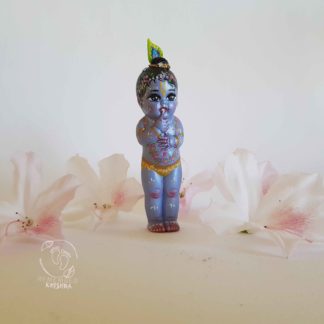 Gopal krishna doll standing among white azaela flowers on a white backdrop wearing a peacock feather and looking like trouble and mischeif