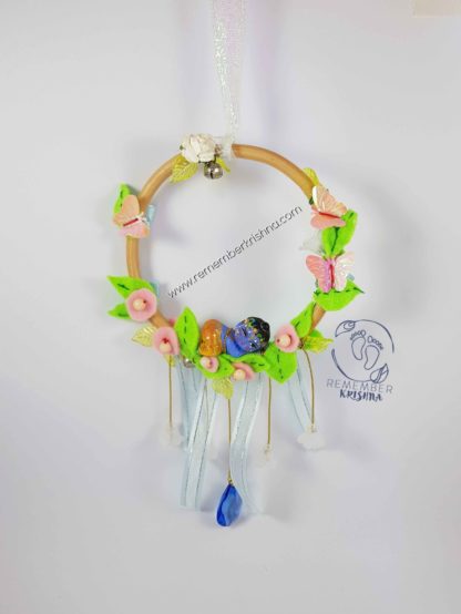 darling baby krsna doll nestled into leaves in a wooden hoop with a hanging blue jewel and sweet suspended flowers, pastel pink and leaves for wind chime which dangles like a dreamcatcher