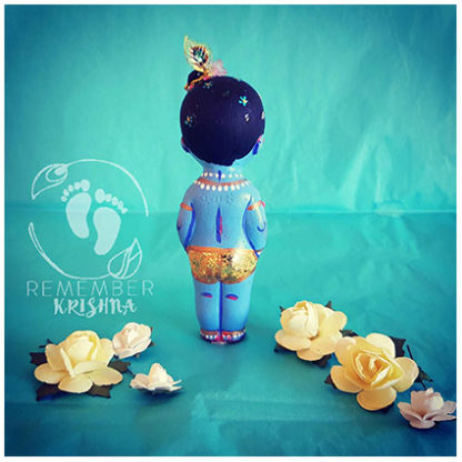 little standing painted krsna doll of hard plastic viewed from behind with chubby knees and peacock feather in his hair and star jewels as well as a turquoise background kahna doll for sale