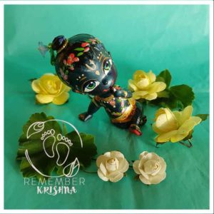 syamasundar Krishna_doll black skinn krsna doll for sale with lotus garland flower crown sitting on a turquoise background with yellow paper flowers around him for kids fun