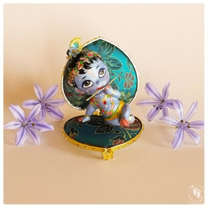 Krishna doll sits on Vyasasana blue heart throne with purple flowers as he plays flute and has peacock feather in hair on pink background for sale