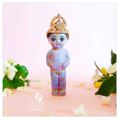 krishna wear beautiful crown from diamonte little krishna deity colored blue on pink background with white orange blossom flowers and petals kahna deity doll for sale