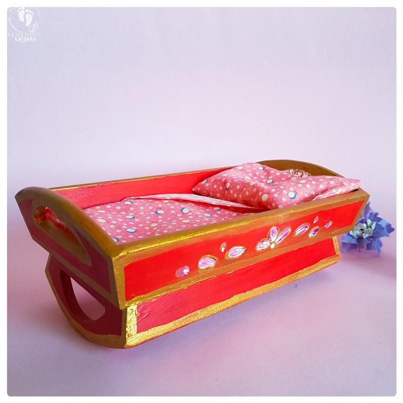 Baby krsna doll blue boy krsna tucked into a wooden rocking cradle painted red with folkstyle art flowers in gold on the sides of the cot and a pink sheet