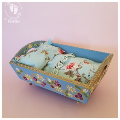 rocking cradle for Krsna dolls painted blue with daisy and blueberry folk art style and blue flowery bedding wooden cradle on pink background