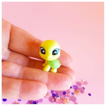 tiny turtle toy, little green turtle being held in hand on a pink background with purple sequins