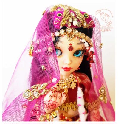 Radhe glancing shyly from behind her veil dressed in red jewels and pink like a bride