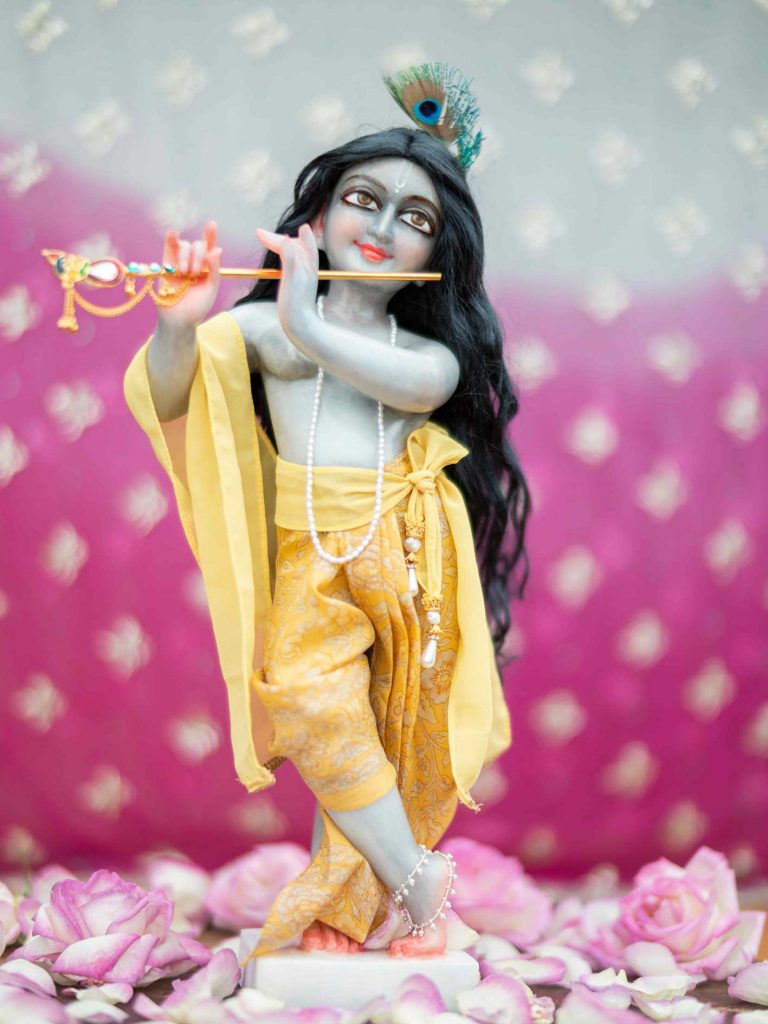 Marble krishna deity stormy blue hue against a pink spotted background wearing a luscious black deity wig hairs for murti and yellow dhoti yellow chaddar yellow sash and dupatta