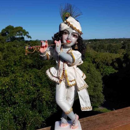 18 inch marble deity krishna painted dusky blue wearing white shringara outfit and mukut on green forest and blue sky backdrop