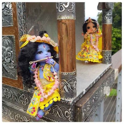 Krishna doll plays his flute sweetly while Radha is in the background krishna wears golden garments and lots fo flowers and sits in a silver and bronze temple radha looks on
