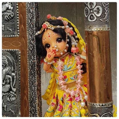 from a silver and bronze temple columns Sri Radharani looks. beautifully decorated poseable ball jointed Radha doll wearing yellow indian gopi dress outfit and lots of pink flowers with pearls and golden jewellery