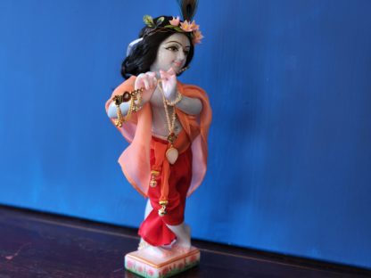 krishna deity 12 inch white marble blue colorwash hue skin dressed in red dhoti murti in three fold bending post with flute peach shawl hair and peacock feather on blue background