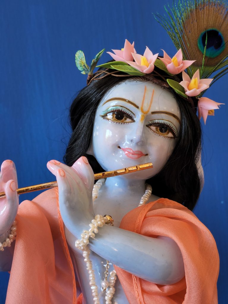 krishna deity 12 inch white marble blue colorwash hue skin dressed in red dhoti murti in three fold bending post with flute peach shawl hair and peacock feather on blue background