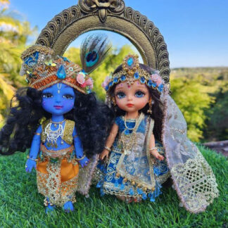 Radha Krishna dolls standing in front of a golden arch and a blue sky background. Radha and Krishna are both dressed in very opulent shringar - Krishna wears a turban and peacock feather as well as an orange dhoti and golden vest. Shri Radhe wears a blue embellished lengha with golden dupatta and special golden beaded crown with flowers and sapphire gems. They are presented as in Vrindavan forest for delight of vaishnava devotees.