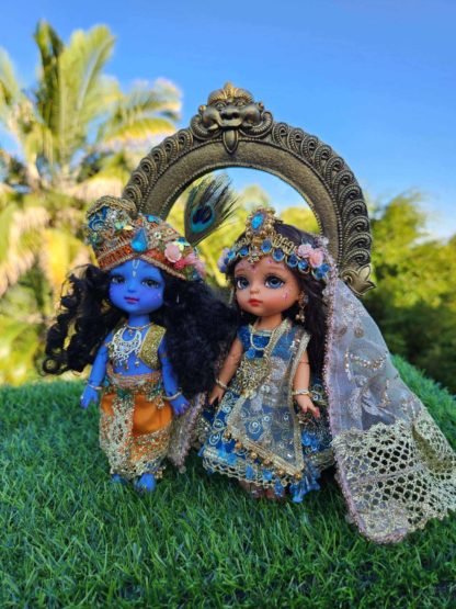 Radha Krishna dolls standing in front of a golden arch and a blue sky background. Radha and Krishna are both dressed in very opulent shringar - Krishna wears a turban and peacock feather as well as an orange dhoti and golden vest. Shri Radhe wears a blue embellished lengha with golden dupatta and special golden beaded crown with flowers and sapphire gems. They are presented as in Vrindavan forest for delight of vaishnava devotees.