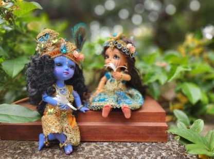 Radha Krishna dolls standing in front of a golden arch and a blue sky background. Radha and Krishna are both dressed in very opulent shringar - Krishna wears a turban and peacock feather as well as an orange dhoti and golden vest. Shri Radhe wears a blue embellished lengha with golden dupatta and special golden beaded crown with flowers and sapphire gems. They are presented as in Vrindavan forest for delight of vaishnava devotees.