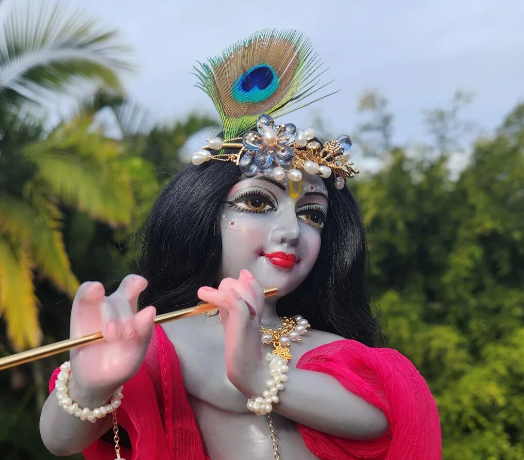 Krishna deity Radharani Krishna stormy blue grey skin against a forest backdround. Krishna wears peacock feather in crown and is dressed in red with pearl bracelets