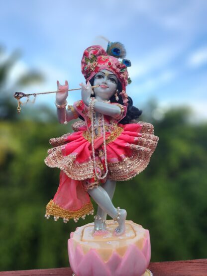 deity of krishna dressed in elaborate pink strawberry inspired shringar playing flute and standing on lotus base