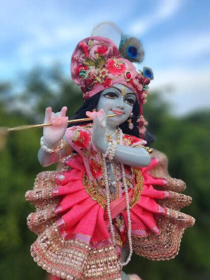 deity of krishna dressed in elaborate pink strawberry inspired shringar playing flute and standing on lotus base