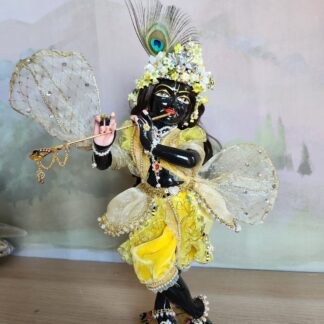 A beautifully adorned, thirteen-inch tall black marble deity of Krishna, known as Sarveshwara Krishna. He is dressed in a fresh bumblebee outfit featuring a rich velvet silk dhoti, a gauzy overskirt decorated with pearls and flowers, and a striped vest with yellow ruffles and diamond trim. His opulent mukut (crown) sparkles with diamonds and is adorned with soft, spring-like flowers. A light, gauzy shawl with subtle diamond glints flutters around Him. A soft, open rose is offered at His feet.