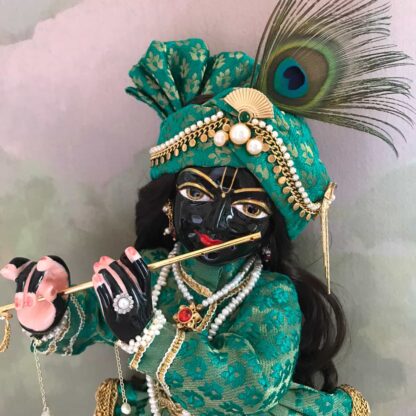 Black marble deity of Krishna wearing a peacock feather for comparison next to the form of the lord dressed in gold and green shringar