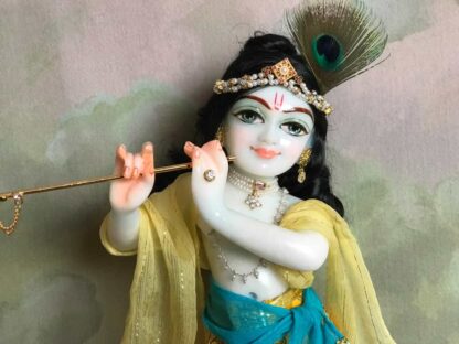 Quality white marble krishna deity hand carved Kamal Nayana weearing soft pink outfit with delicate pink hands in a pink turban and a peacock feather with delicate pearl and gold jewelery 12inch tall deity of Krishna shown holding flute. deity pictured in slight side profile focus on face and peacock feather with jeweled crown