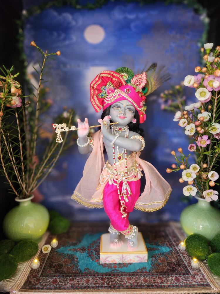Marble deity of Krishna dressed in elaborate pink Shringar with pearl beading, embroidery turban covered in stonework and pearls with peacock feathers and flowers and a long sleeve jacket heavily embroidered. Deity is ornamented with pearl anklets, pearl bracelets, arm cuffs, long three strand necklaces with ruby red stones. Deity of Krishna plays the flute