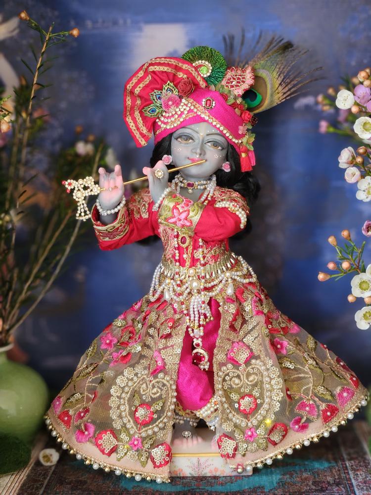 Marble deity of Krishna dressed in elaborate pink Shringar with pearl beading, embroidery turban covered in stonework and pearls with peacock feathers and flowers and a long sleeve jacket heavily embroidered. Deity is ornamented with pearl anklets, pearl bracelets, arm cuffs, long three strand necklaces with ruby red stones. Deity of Krishna plays the flute
