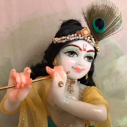 Quality white marble krishna deity hand carved Kamal Nayana weearing soft pink outfit with delicate pink hands in a pink turban and a peacock feather with delicate pearl and gold jewelery 12inch tall deity of Krishna shown holding flute. deity pictured in slight side profile focus on face and peacock feather with jeweled crown