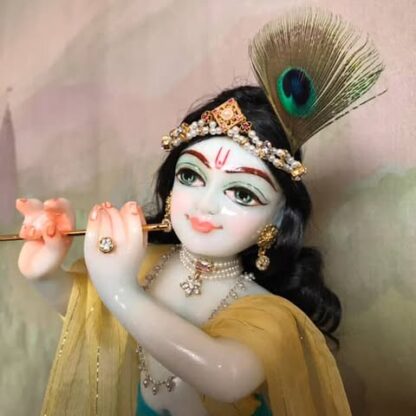 Quality white marble krishna deity hand carved Kamal Nayana weearing soft pink outfit with delicate pink hands in a pink turban and a peacock feather with delicate pearl and gold jewelery 12inch tall deity of Krishna shown holding flute. deity pictured in slight side profile focus on face and peacock feather with jeweled crown