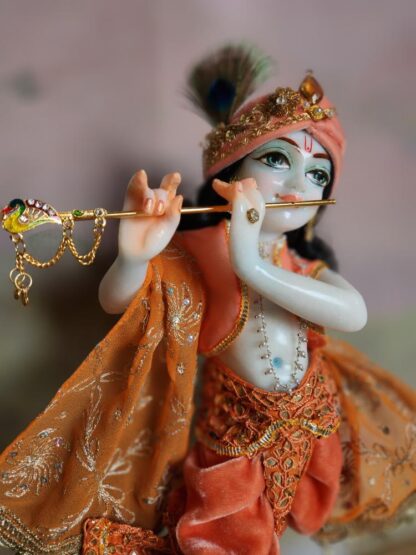 BalaGopal deity Quality white marble krishna deity hand carved Kamal Nayana weearing soft pink outfit with delicate pink hands in a pink turban and a peacock feather with delicate pearl and gold jewelery 12inch tall deity of Krishna shown holding flute. deity pictured in slight side profile focus on face and peacock feather with apricot and peach gorgeous dhoti