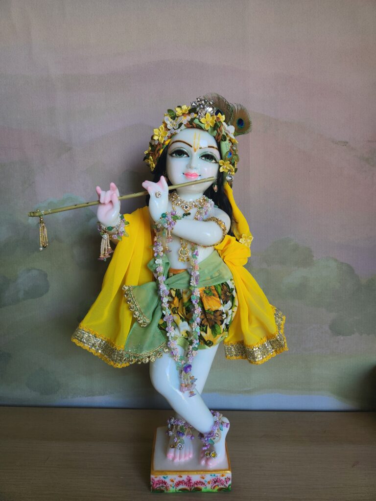 A marble Gopal Krishna deity wearing a vibrant outfit with sunflower-print shorts, a mossy green sash, and a bright yellow shawl.