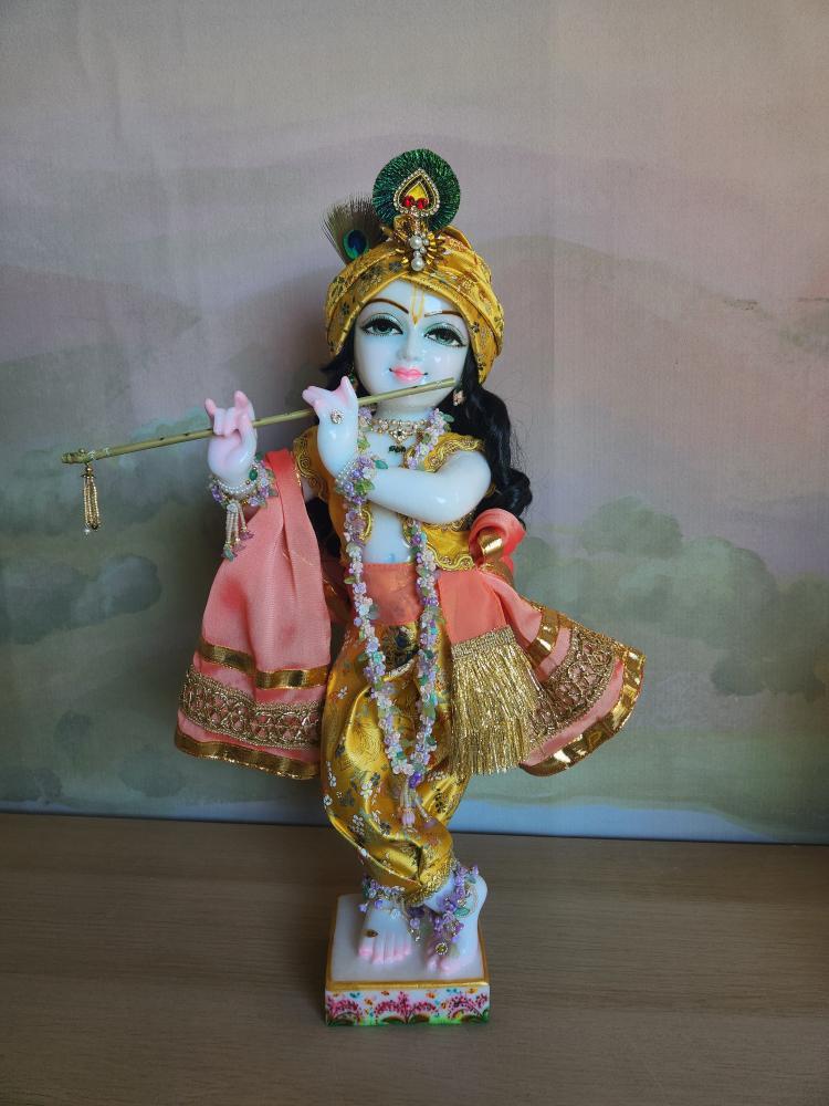 A marble Gopal Krishna deity in full harem pants of silk brocade, a peach sash, a yellow vest with gold braid, and a turban.