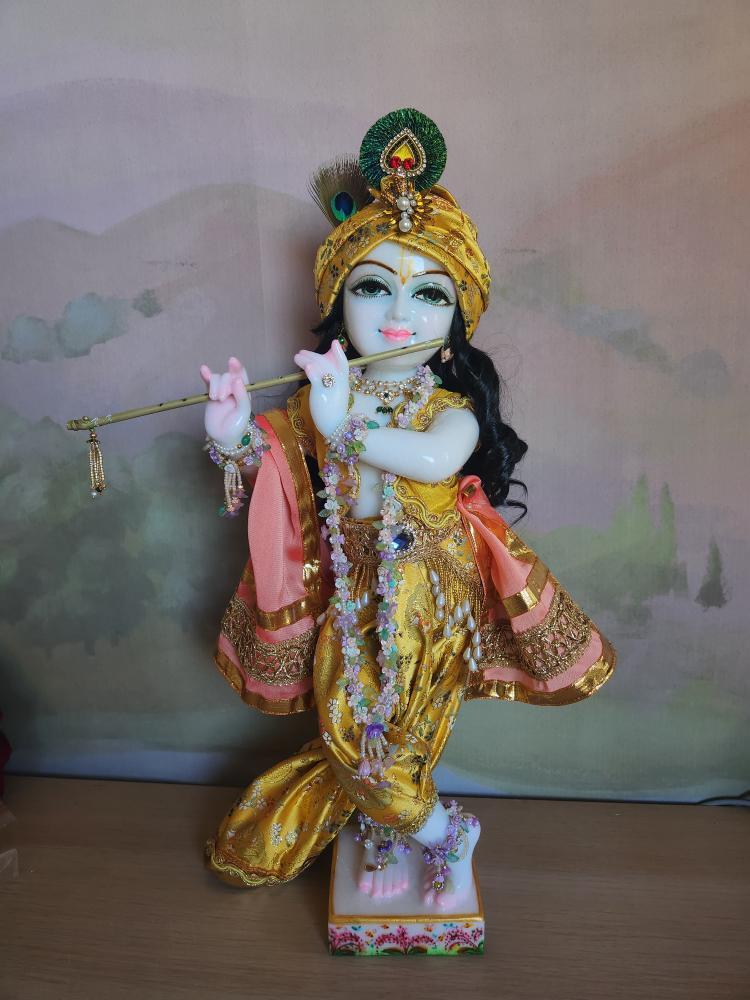 A marble Gopal Krishna deity in full harem pants of silk brocade, a peach sash, a yellow vest with gold braid, and a turban.