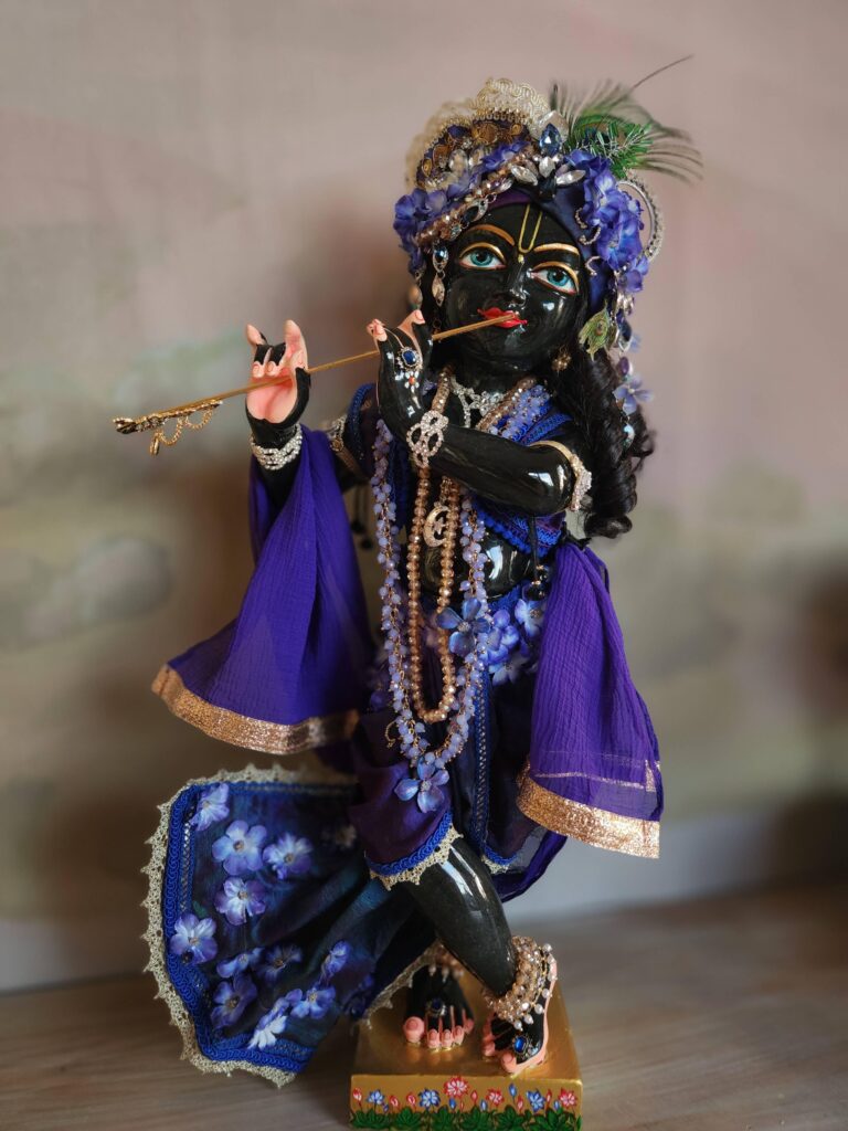 Syamasundar black marble Krishna deity dressed in indigo blue hues with flower outfit and shown playing flute against a sunrise