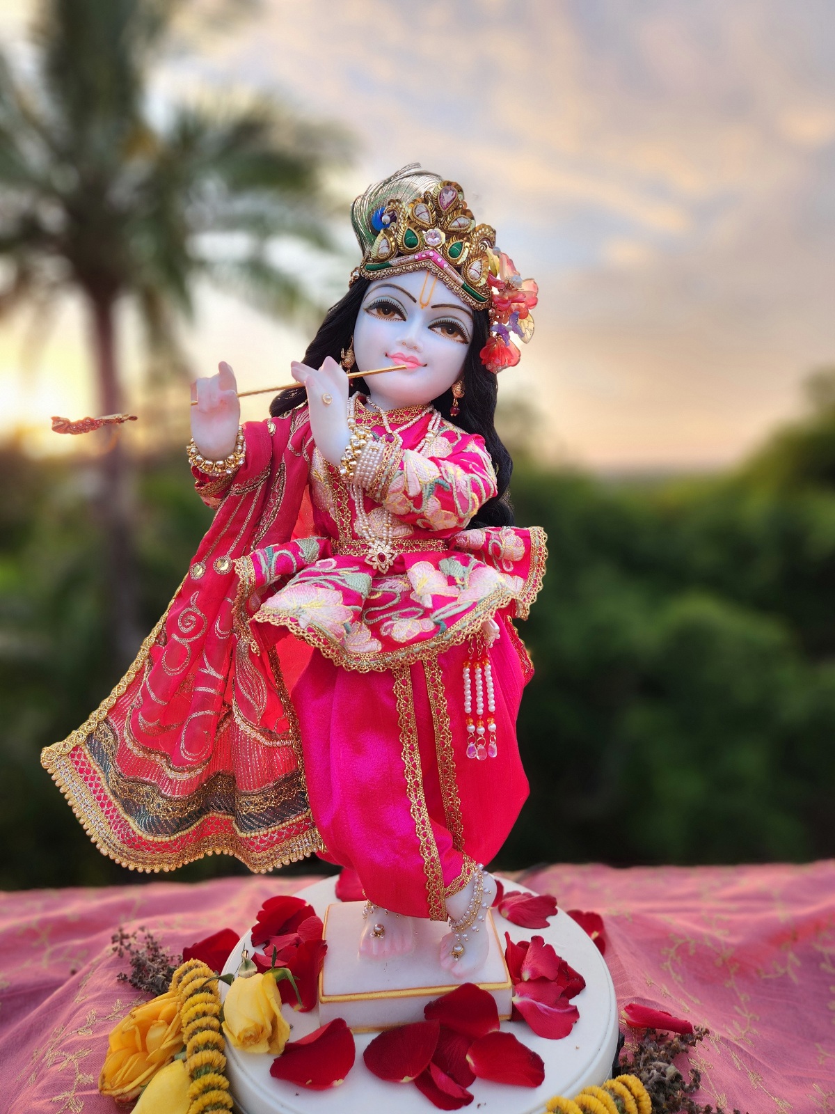 Full view of the unique Krishna deity, named Vrindavanchandra, standing in the classic three-fold bending pose. His moonlit white marble form stands 18-inches tall, radiating an ethereal beauty. He is gorgeously dressed in a couture hot pink outfit of full pants, jacket with lotus embroidery, and a long gold embroidered shawl. He wears a golden crown and pink flowers in his hair. Behind the deity is a nature landscape of greenery and a early morning sunrise.