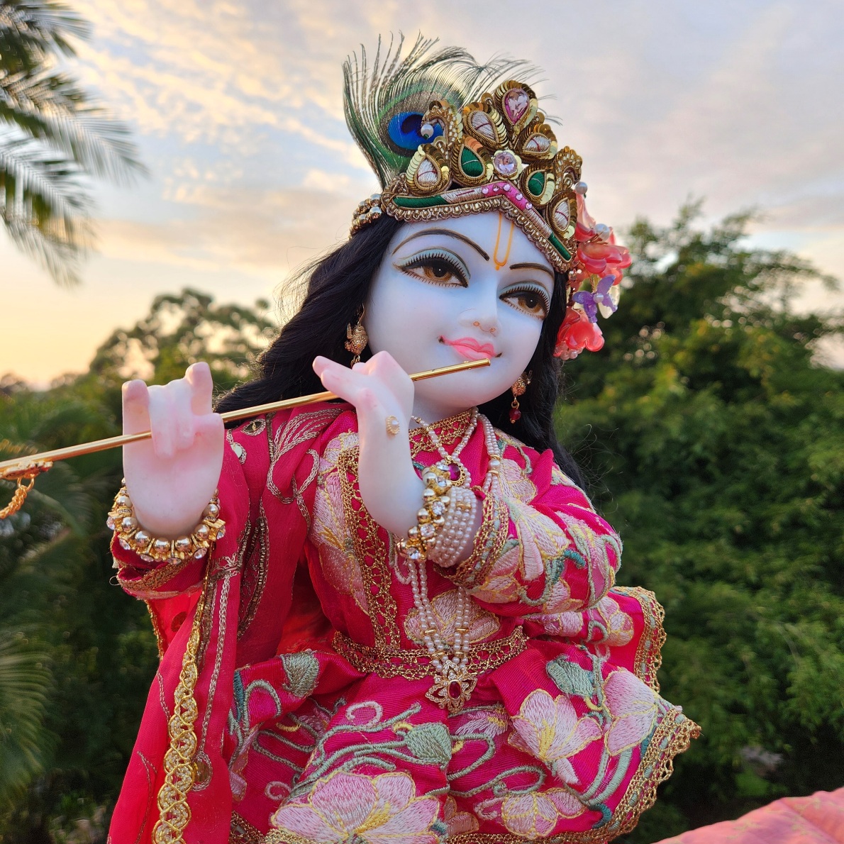 Close-up side view of a unique Krishna deity. Dressed in pink embroidered long sleeve jacket with a full lotus embroidered short peplum, and forest in the background along with a sunrise in the sky. This 18-inch marble form has a moonlit complexion, large smiling eyes, and a mild, gentle smile. He is wearing a black waist-length wig, blue and pearl jewelry, and a natural peacock feather.