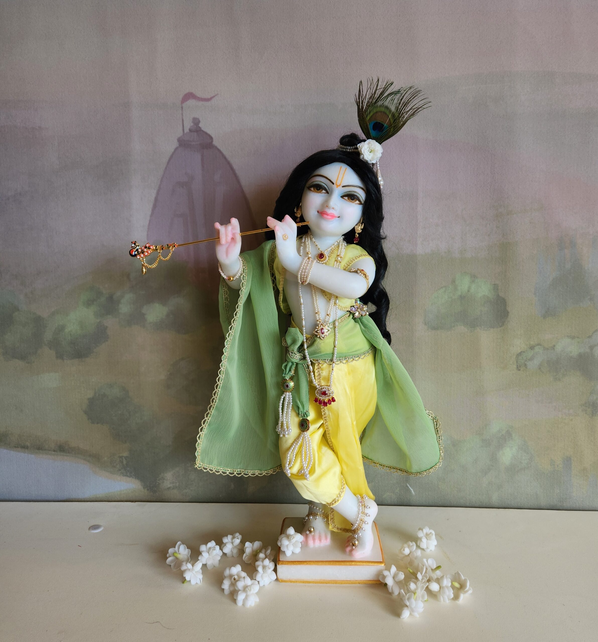 marble deity of krishna painted a soft delicate blue hue wearing flower garlands and pearl jewellery deity wearing yellow and green with pearl tassels on ends of sash