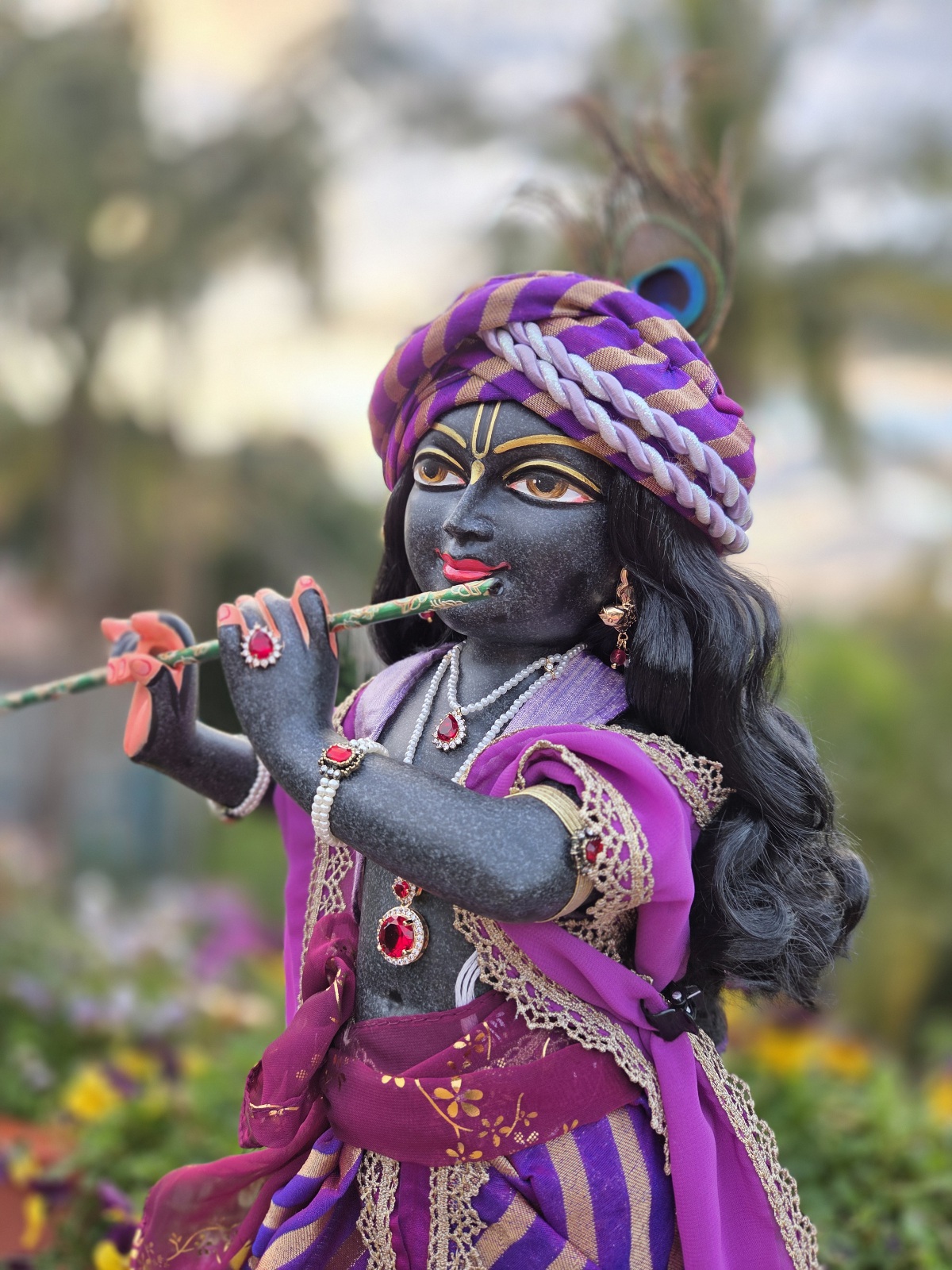 Luxurious black Marble deity of Gopinath Krishna standing with flute to His nectarian lips. Dressed in a purple shringar with gorgeous silks, chiffons, gold lace and brocade. Clothed in a dhoti, short vest, chaddar and pagri, Gopinath a hand-carved black marble Krishna deity, 18 inches tall, with 7 outfits & flower-jewel shringar included. One of a kind — free shipping. Photographed against a sky and garden backdrop