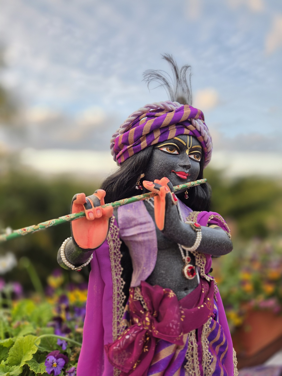 Luxurious black Marble deity of Gopinath Krishna standing with flute to His nectarian lips. Dressed in a purple shringar with gorgeous silks, chiffons, gold lace and brocade. Clothed in a dhoti, short vest, chaddar and pagri, Gopinath a hand-carved black marble Krishna deity, 18 inches tall, with 7 outfits & flower-jewel shringar included. One of a kind — free shipping. Photographed against a sky and garden backdrop