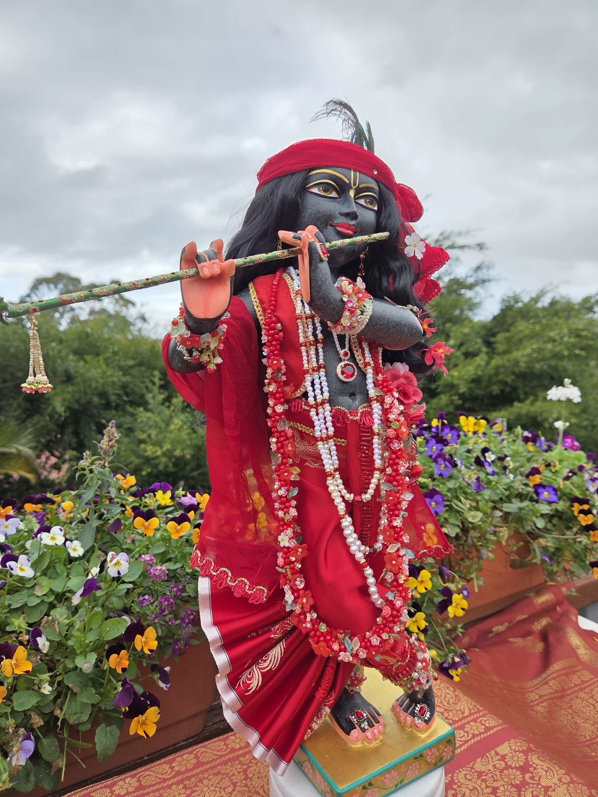 side view of hand carved deity of Krishna black marble 18inches tall photo standing with ornate red shringar against a forest backdrop with flowers and sky