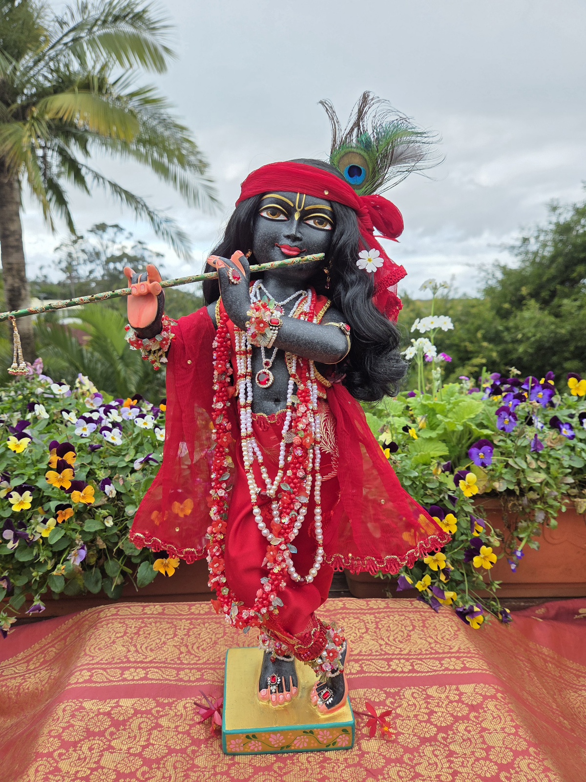 hand carved deity of Krishna black marble 18inches tall photo standing with ornate red shringar against a forest backdrop with flowers and sky
