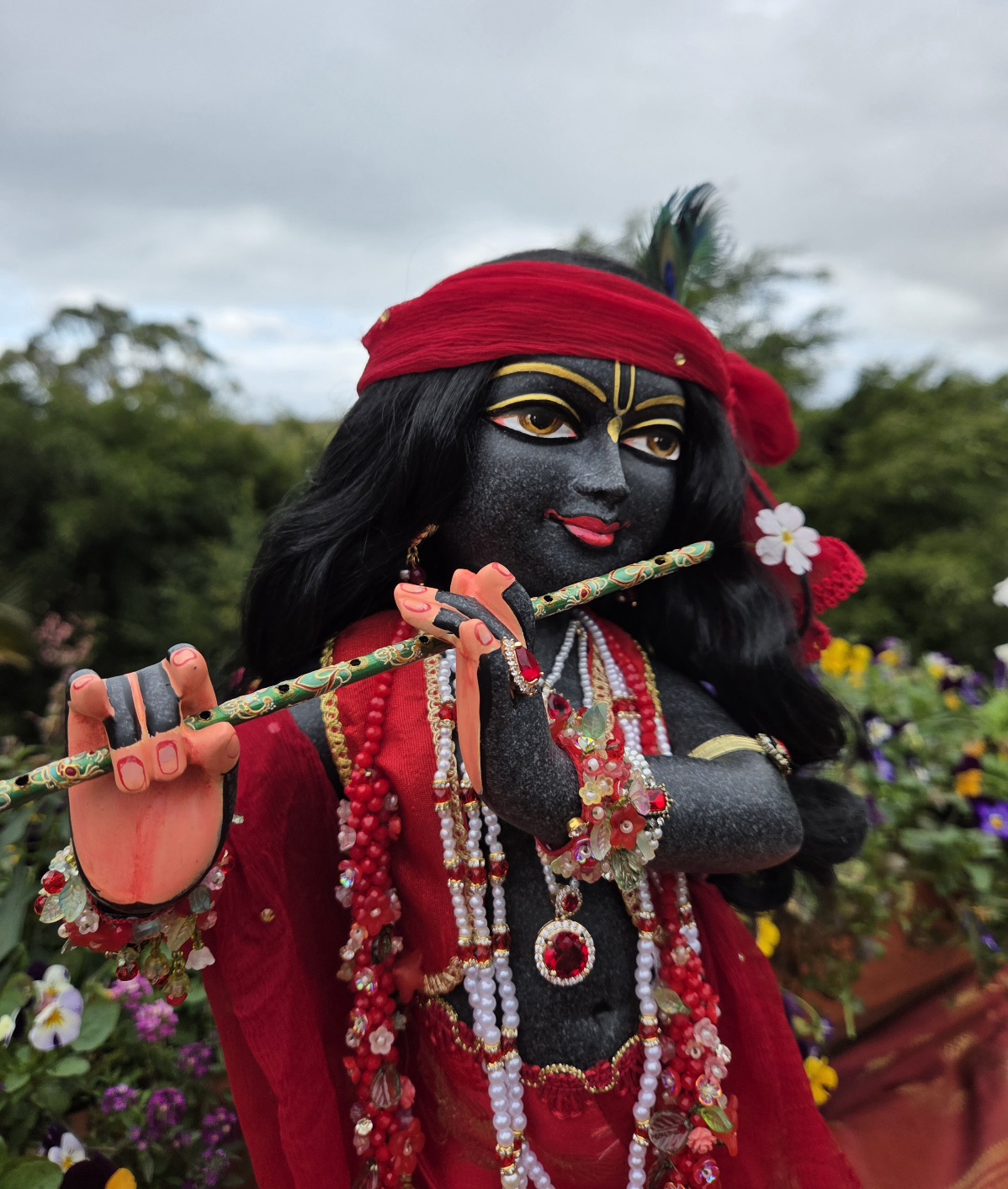close up hand carved deity of Krishna black marble 18inches tall photo standing with ornate red shringar against a forest backdrop with flowers and sky
