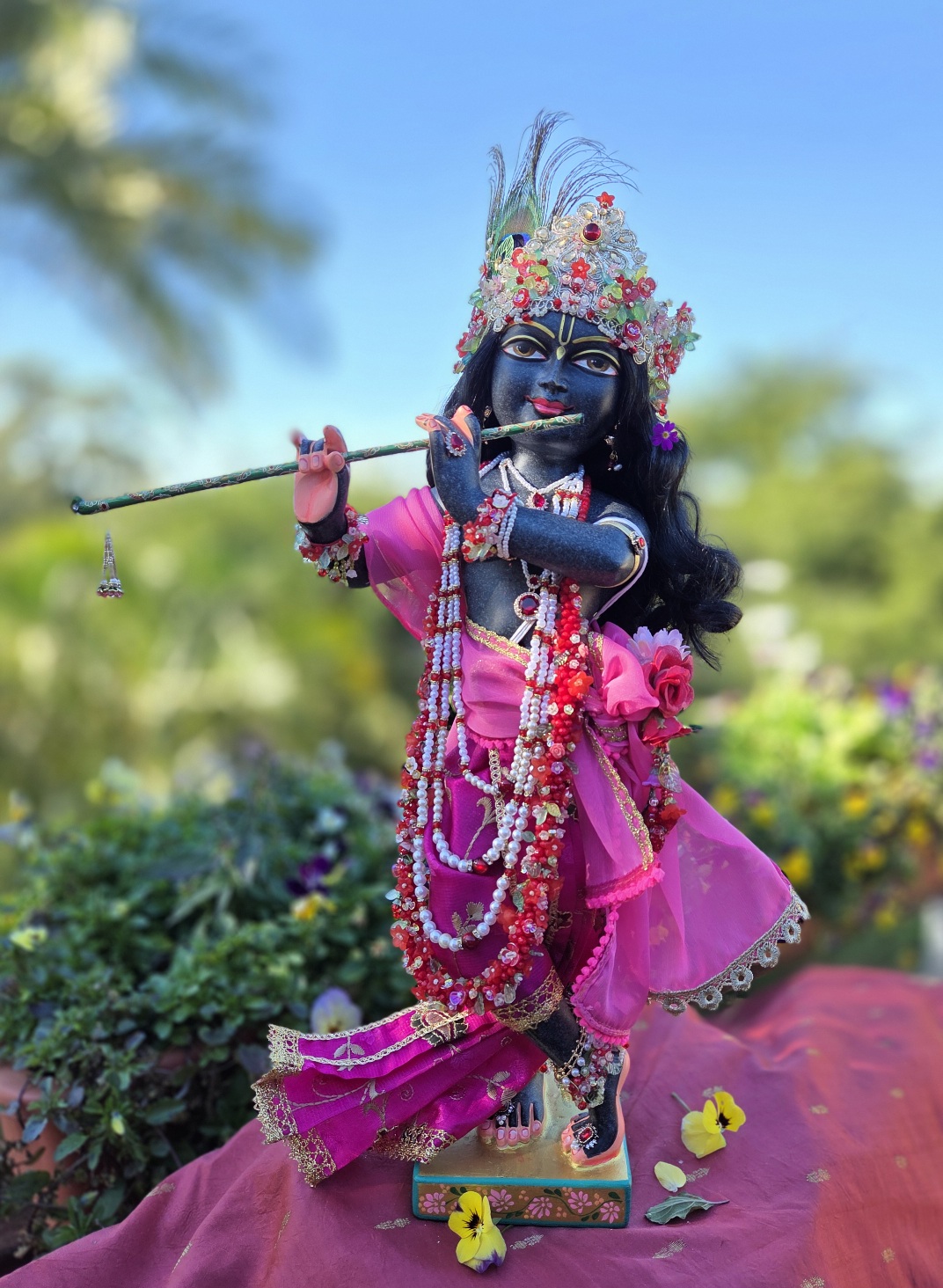 Krishna deity, 18inch tall black marble wearing pink clothes and pearls. His flower crown is very pretty and he is against a forest scene