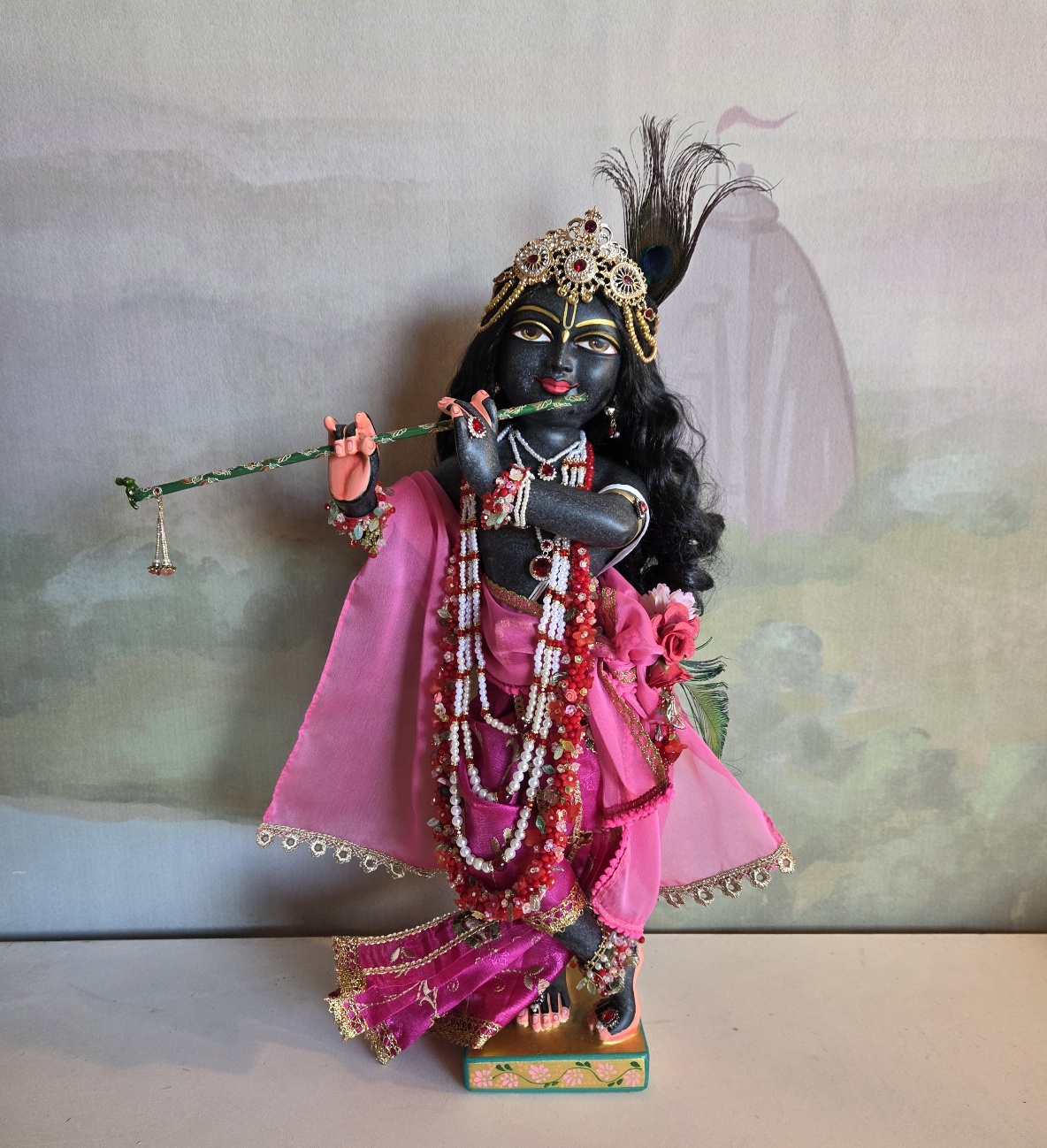 Krishna deity carved from black marble pictured against a watercolor vrindavan backdrop. Deity dressed in pink garments full view