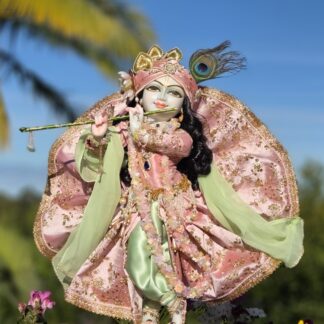 Full view of this 18inch tall white marble Krishna deity. Decoratively arrayed in a soft pink outfit over pistachio green pants deity of Krishna against a blue sky