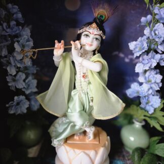 deity of Krishna named Balagopal carved in white marble with a mild blue colorwash. standing on a lotus carved base in a dark blue altar, the deity wears a mint colored shringar