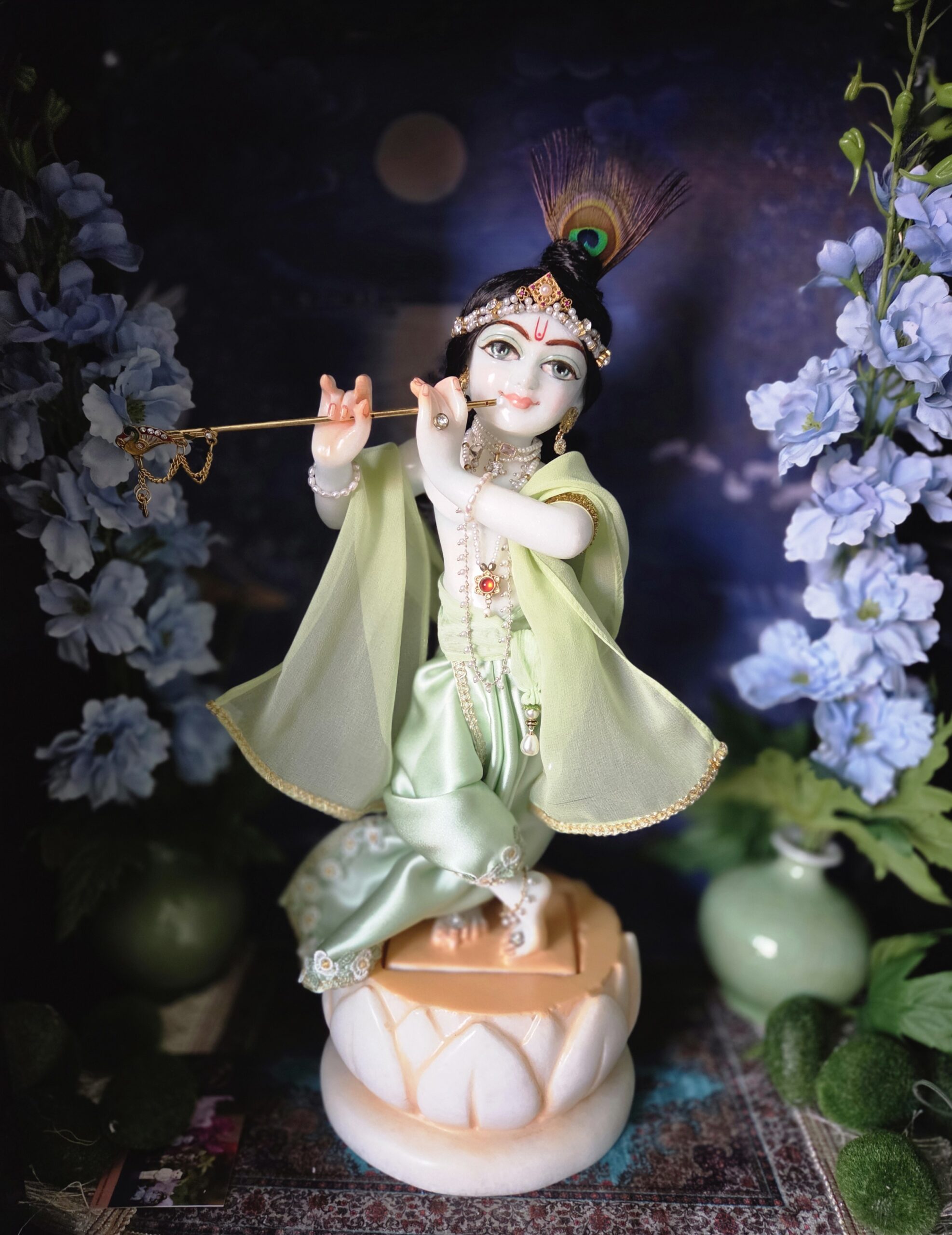 deity of Krishna named Balagopal carved in white marble with a mild blue colorwash. standing on a lotus carved base in a dark blue altar, the deity wears a mint colored shringar