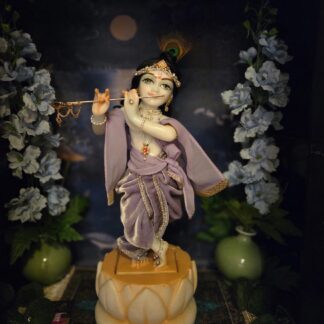 deity of Krishna named Balagopal carved in white marble with a mild blue colorwash. standing on a lotus carved base in a dark blue altar, the deity wears a lavender velvet colored shringar