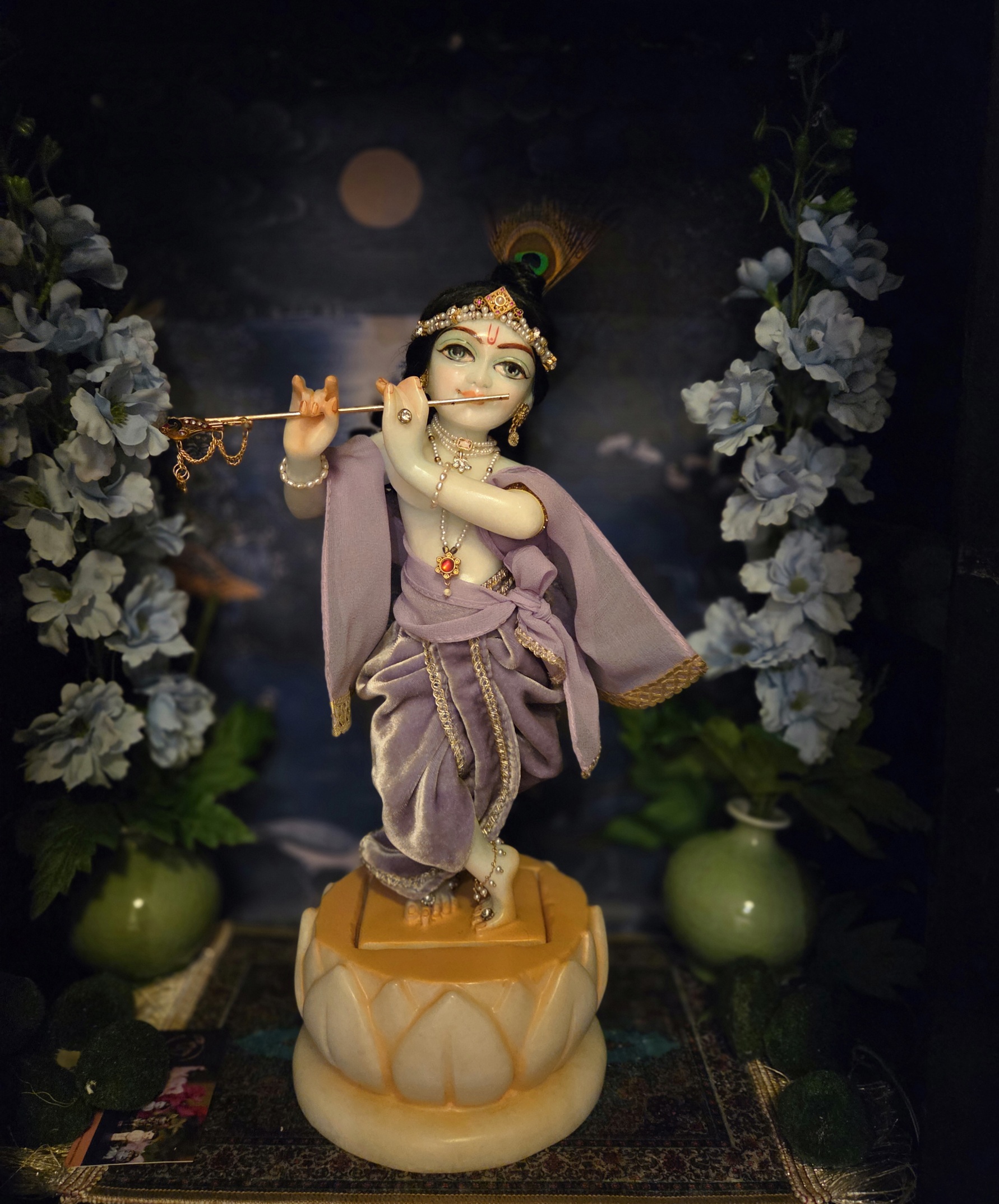 deity of Krishna named Balagopal carved in white marble with a mild blue colorwash. standing on a lotus carved base in a dark blue altar, the deity wears a lavender velvet colored shringar